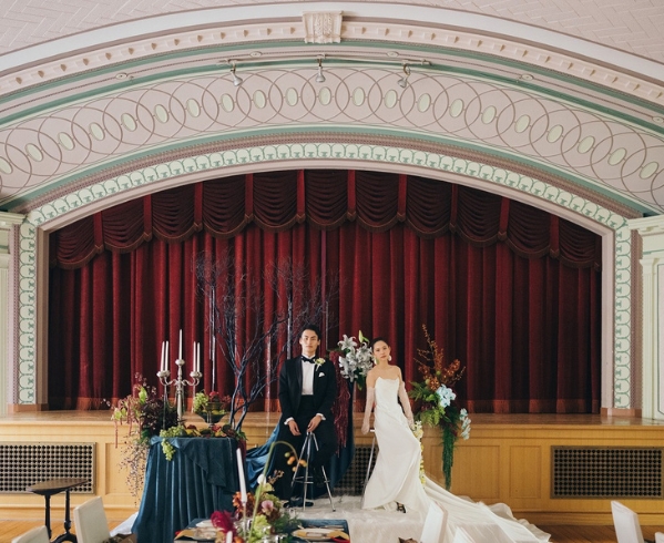 劇場のステージのような空間で、装花に囲まれて座る新郎新婦